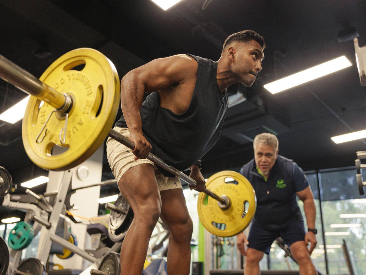 A gym member pulls up a barbell as a trainer watches on