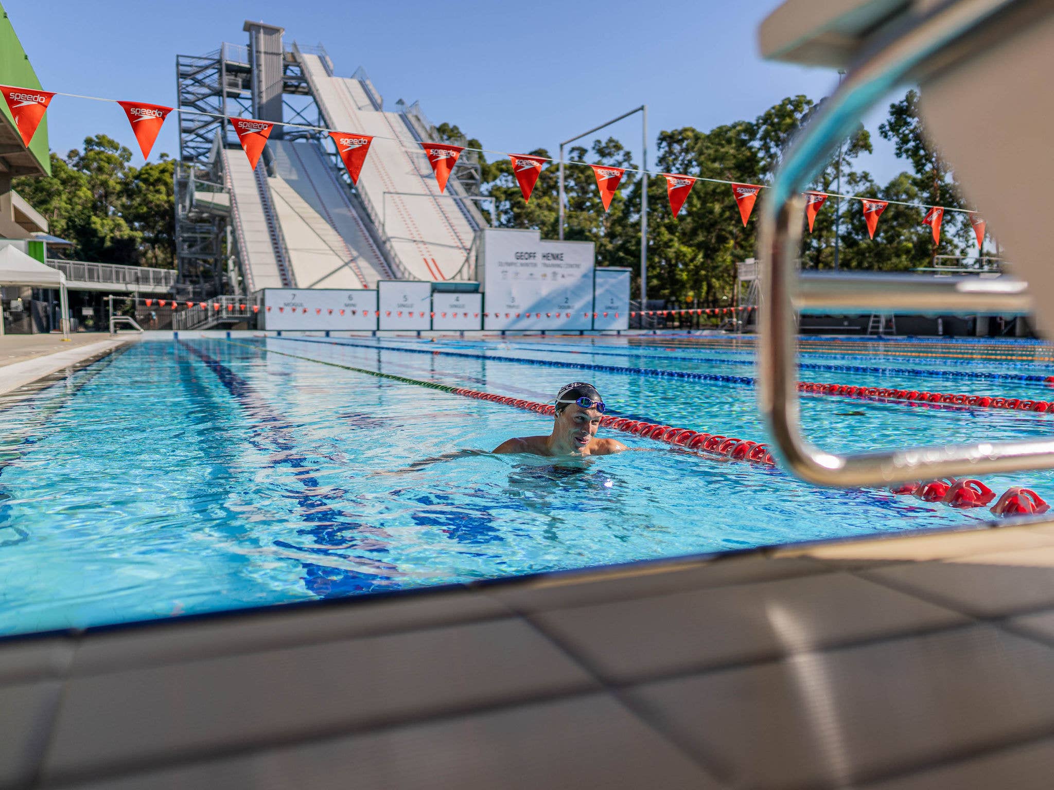 A swimmer floots near the diving block with pool flags above and ski jump in the background